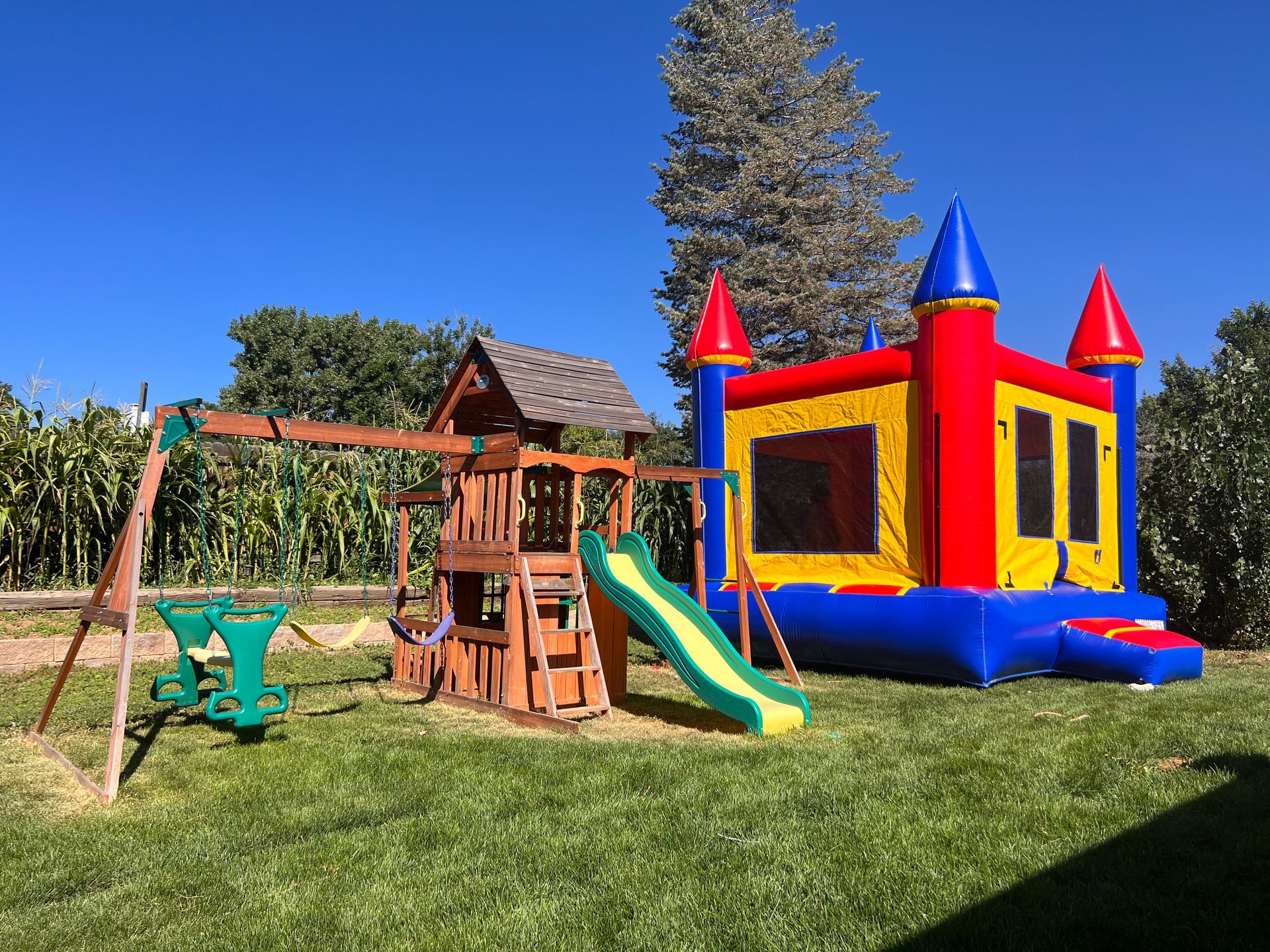 A backyard with a wooden playset, a bounce house, and a swing set on a sunny day.