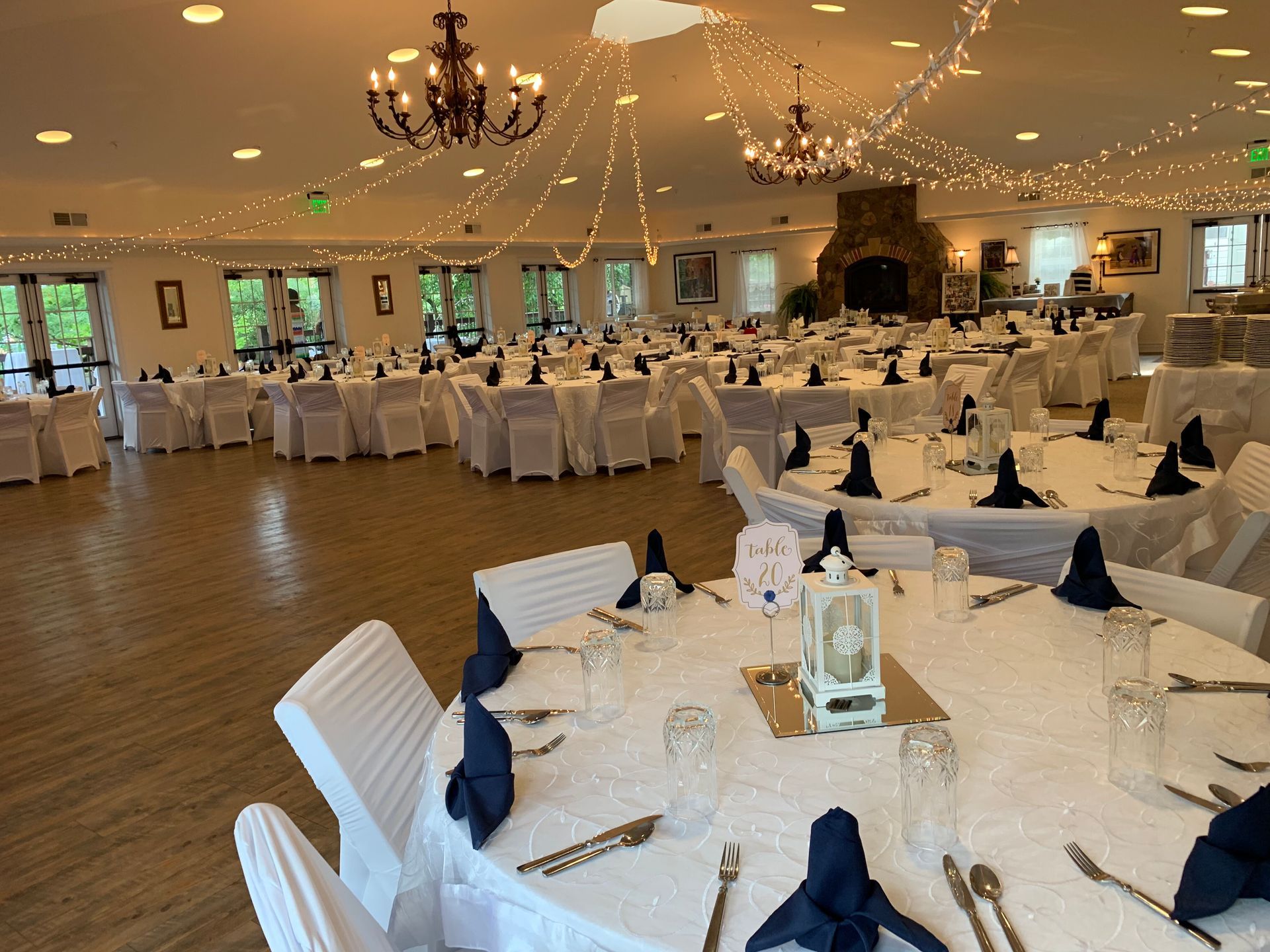 Wedding reception hall with round tables set for guests. White linens, navy napkins, and overhead string lights.