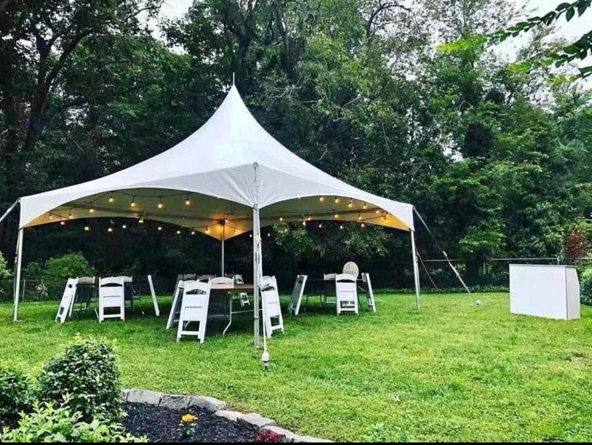 White tent with string lights in a backyard, set for a party with tables and chairs on the grass.