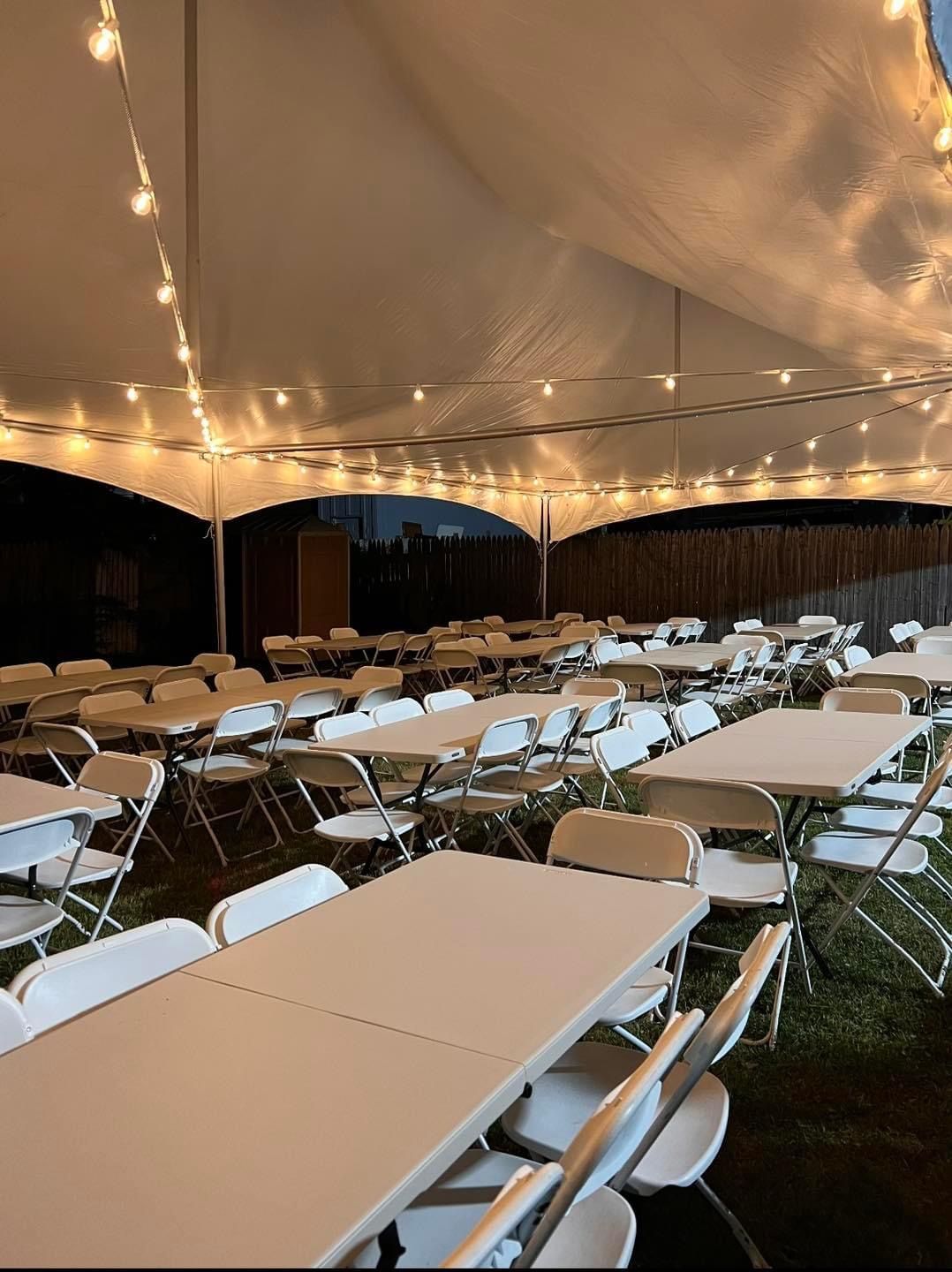 White tables and chairs set up under a tent, strung with lights, outdoors at night.