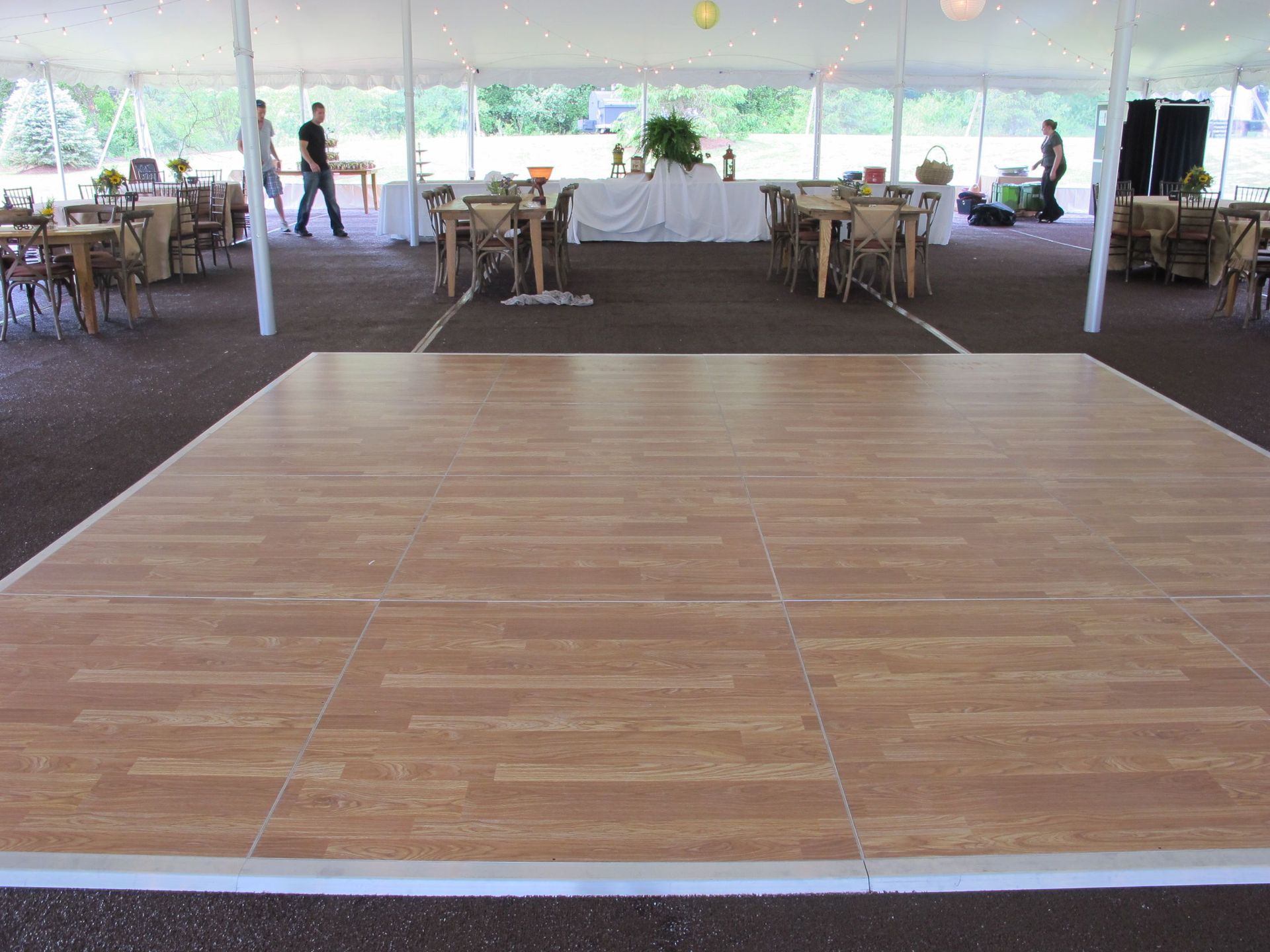 Wooden dance floor in a tent set up for an event, with tables and people in the background.