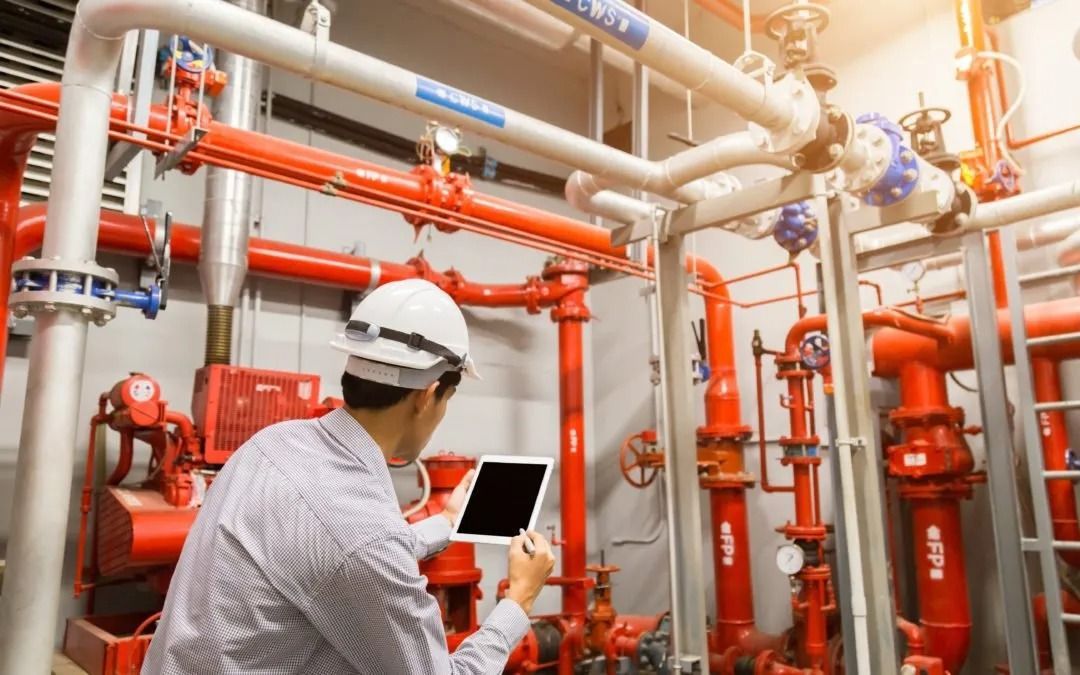 Ingeniero con casco inspeccionando tuberías y válvulas rojas con tableta en un entorno industrial.