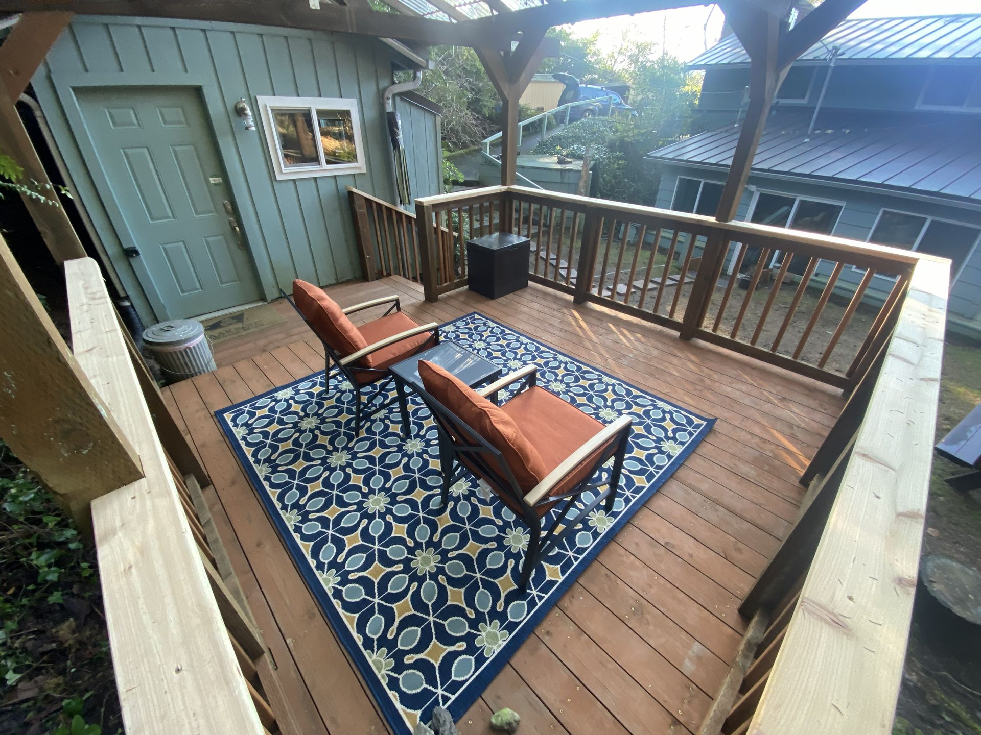 Wooden deck with two chairs, an area rug, and a small table. Green building in the background.