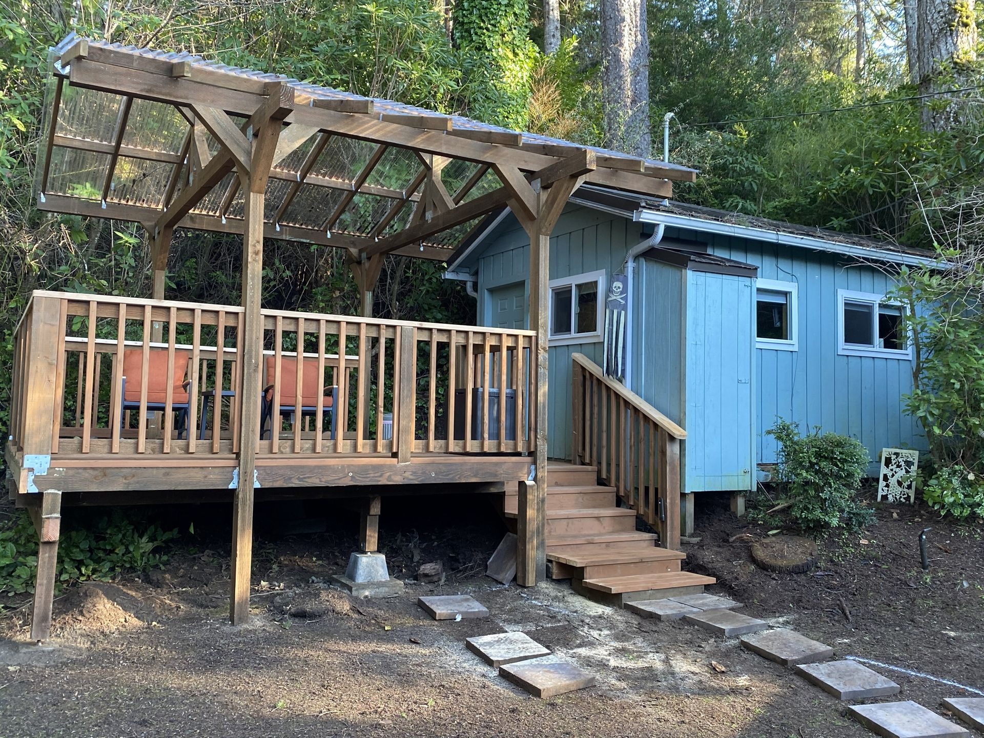 Blue cabin with a wooden deck and pergola in a wooded setting; stone path leads to steps.
