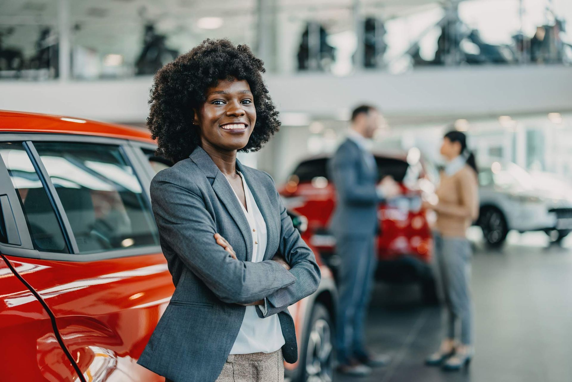 Woman in a car showroom with arms crossed with two people in the background. Woman in a car showroom with arms crossed with two people in the background.