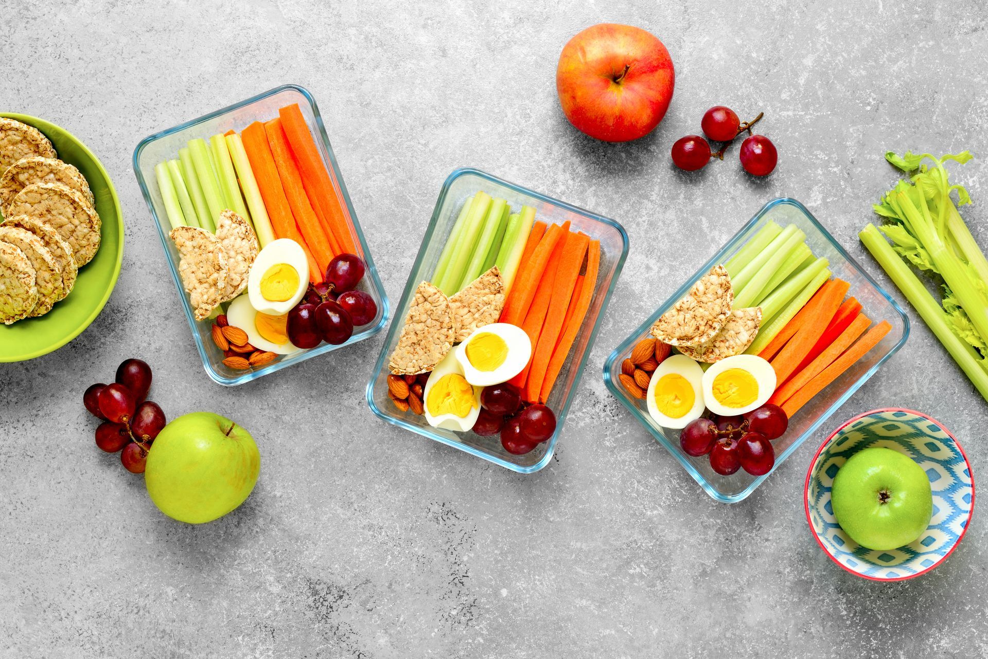 Three glass containers filled with carrots , celery , eggs , grapes , apples and crackers.