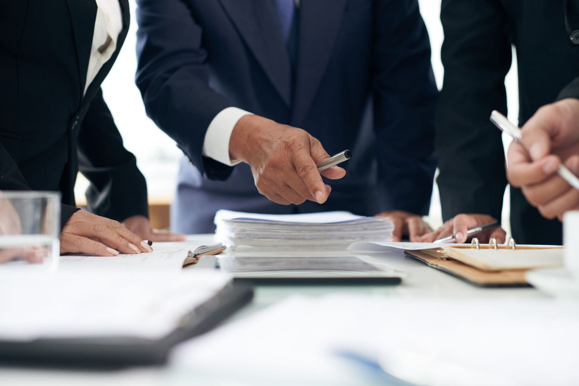 People in suits pointing at documents on a table during a meeting.