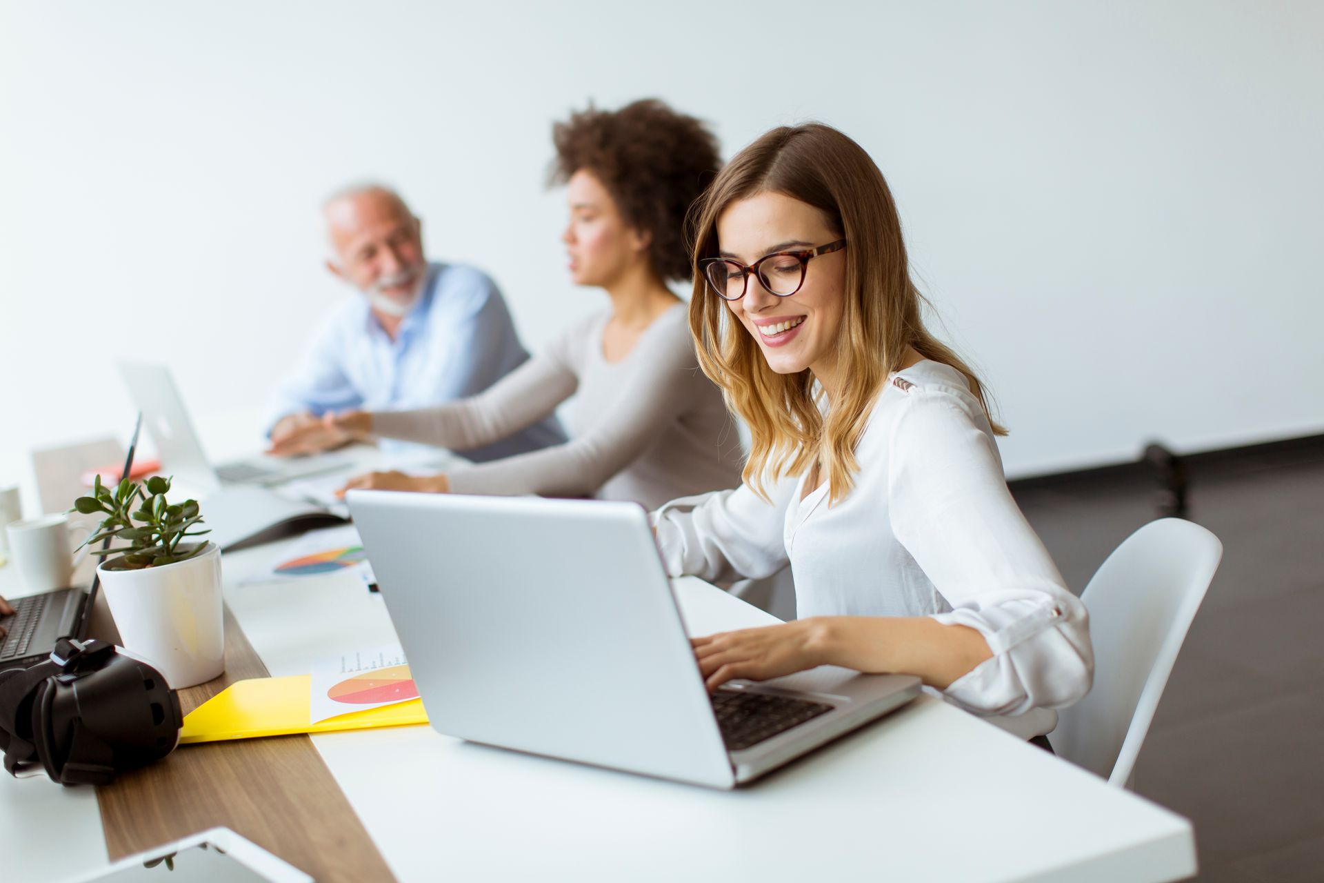 Woman with glasses smiles while typing on a laptop at a white desk; two colleagues in background.