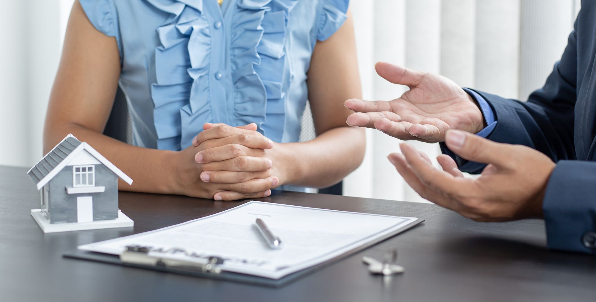 A real estate agent gestures towards a woman, discussing paperwork with a house model and keys on a table.