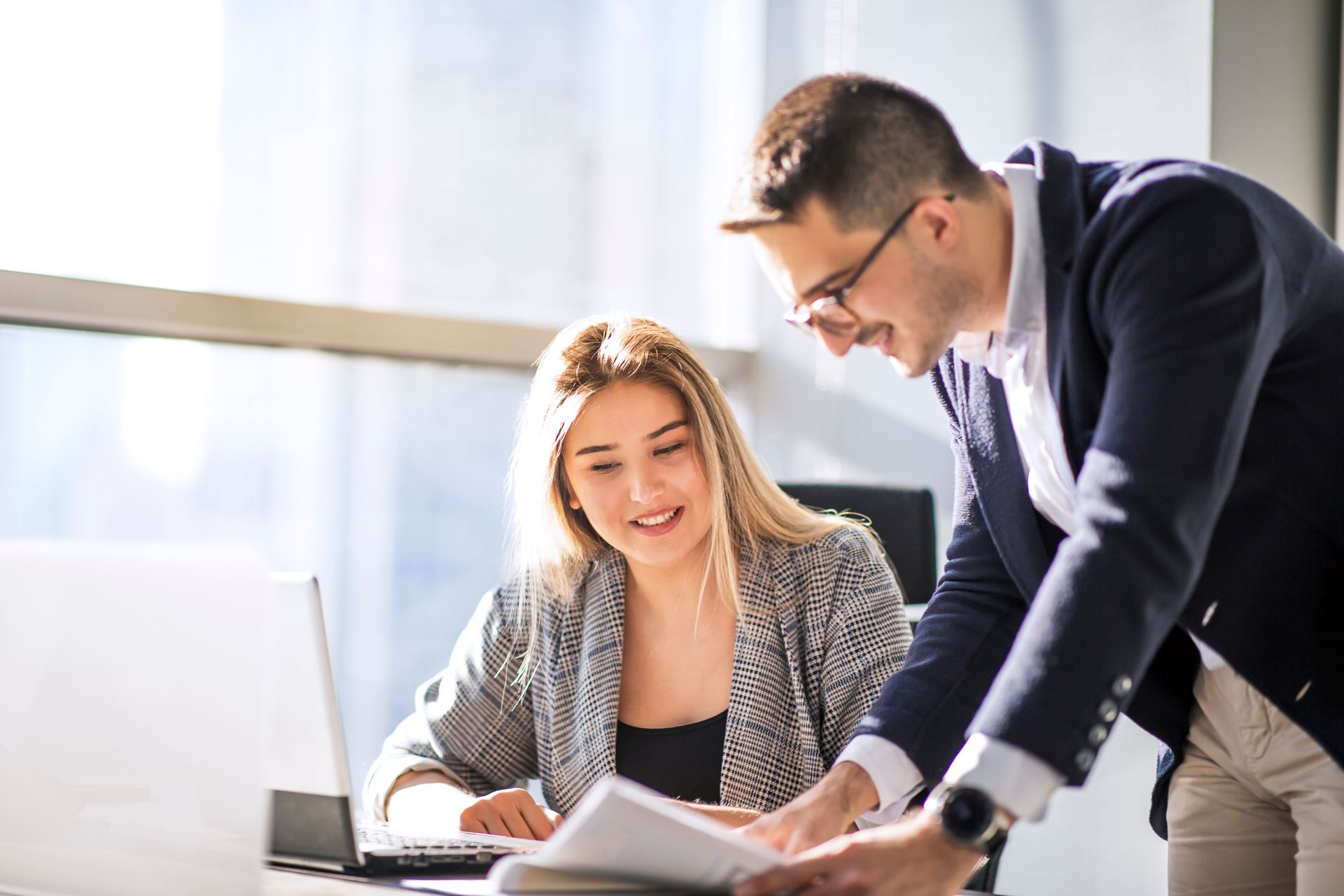 Woman and man collaborating at a desk, reviewing documents near a laptop in an office setting.