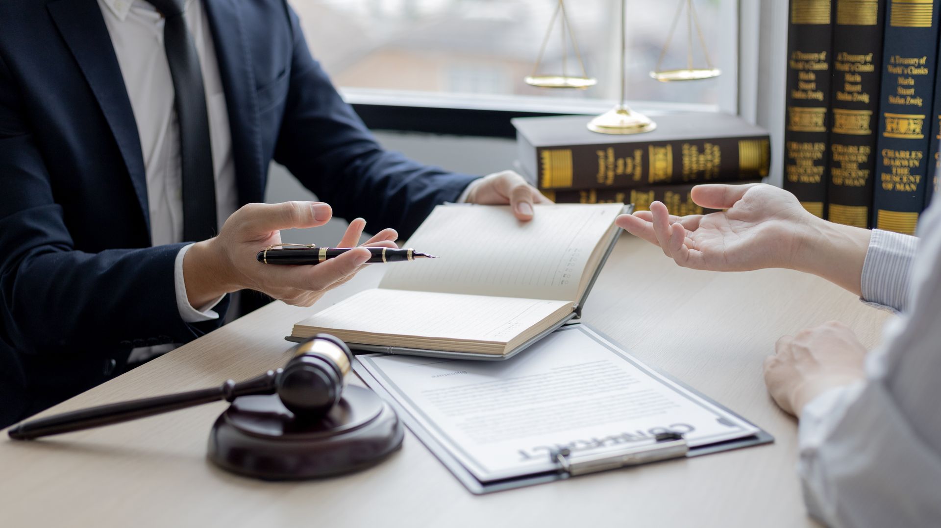 Lawyer discussing document with client; gavel, scales, and law books on the desk.