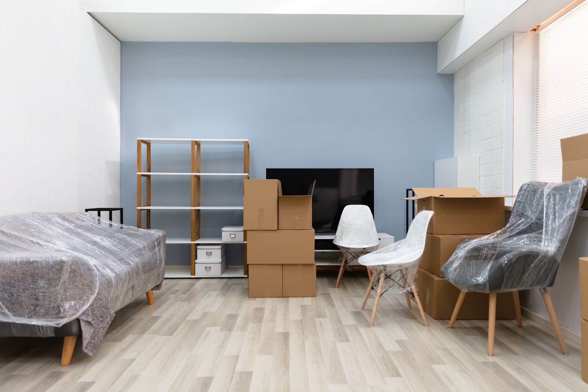 Two specialists in blue overalls are performing a furniture removal service, moving a sofa. View of the interior of an apartment with packed furniture and boxes, and a couch wrapped in plastic.