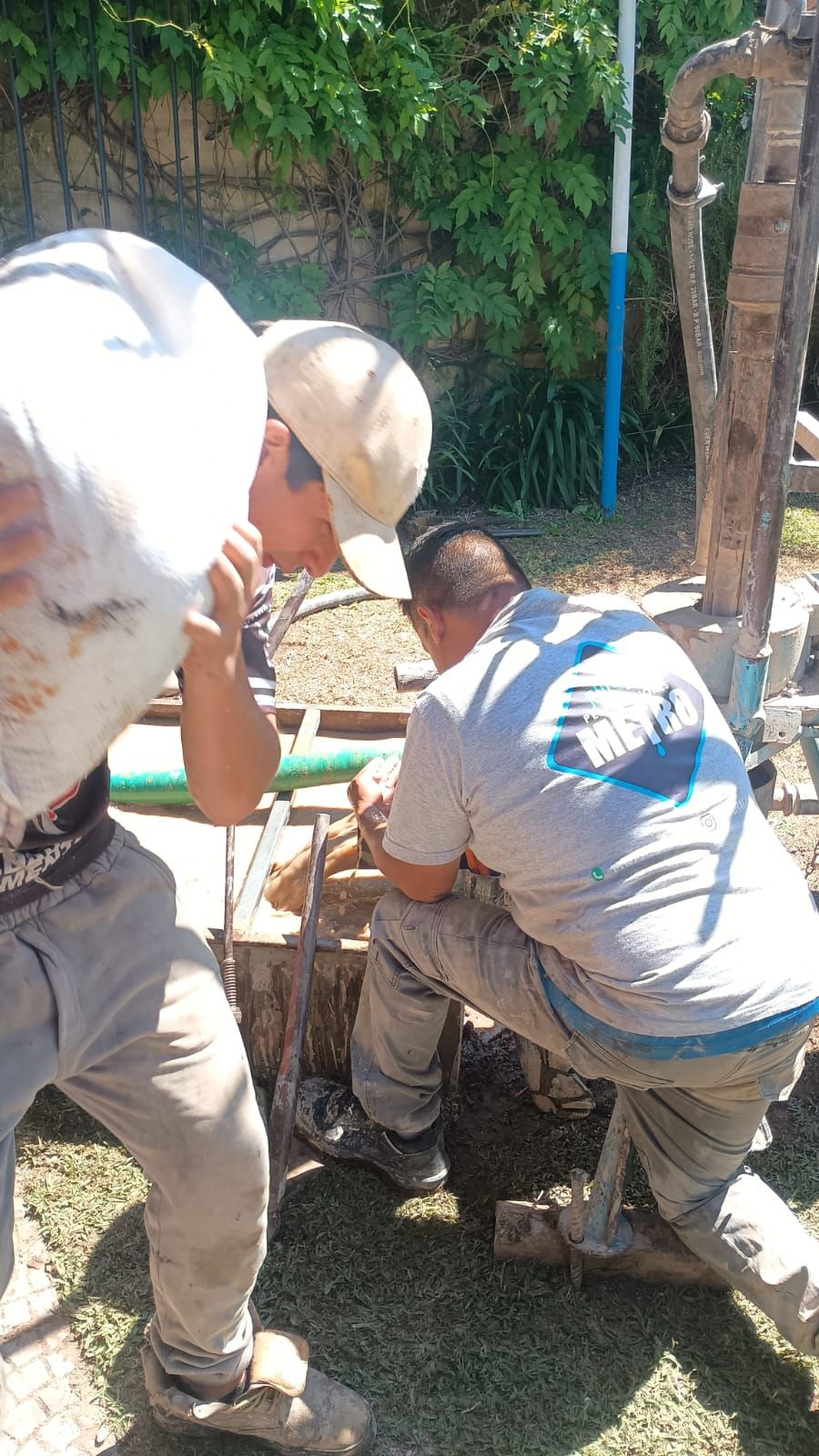 Dos trabajadores con cascos blancos reparan tuberías al aire libre junto a una estructura de hormigón.