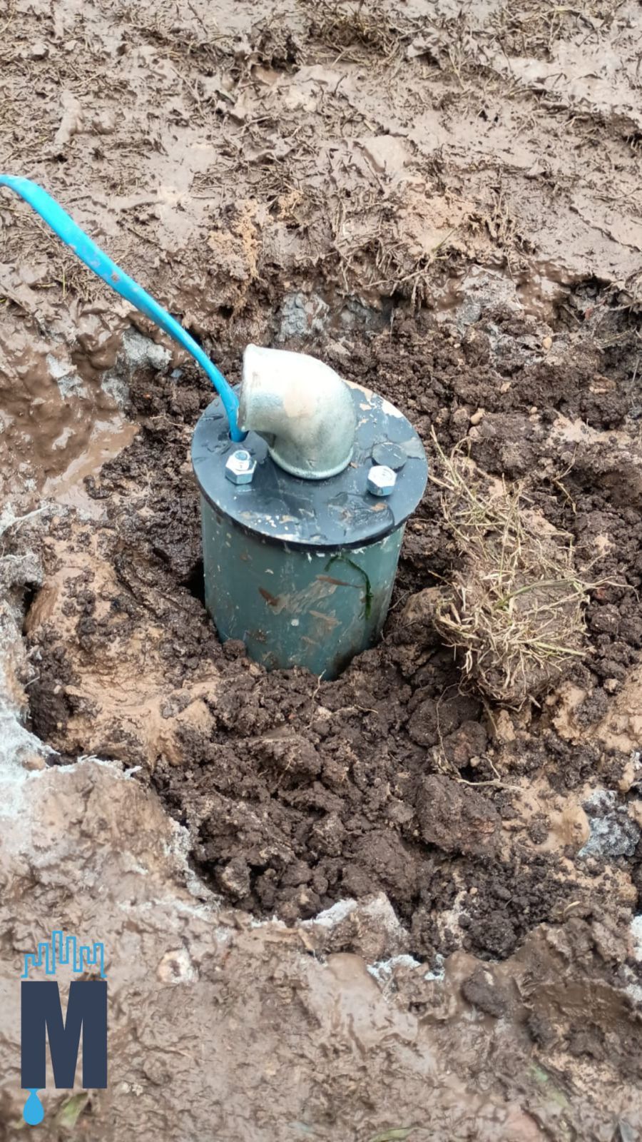 Tubería de servicios públicos verde enterrada con tapa blanca y cable azul que emerge del terreno fangoso.