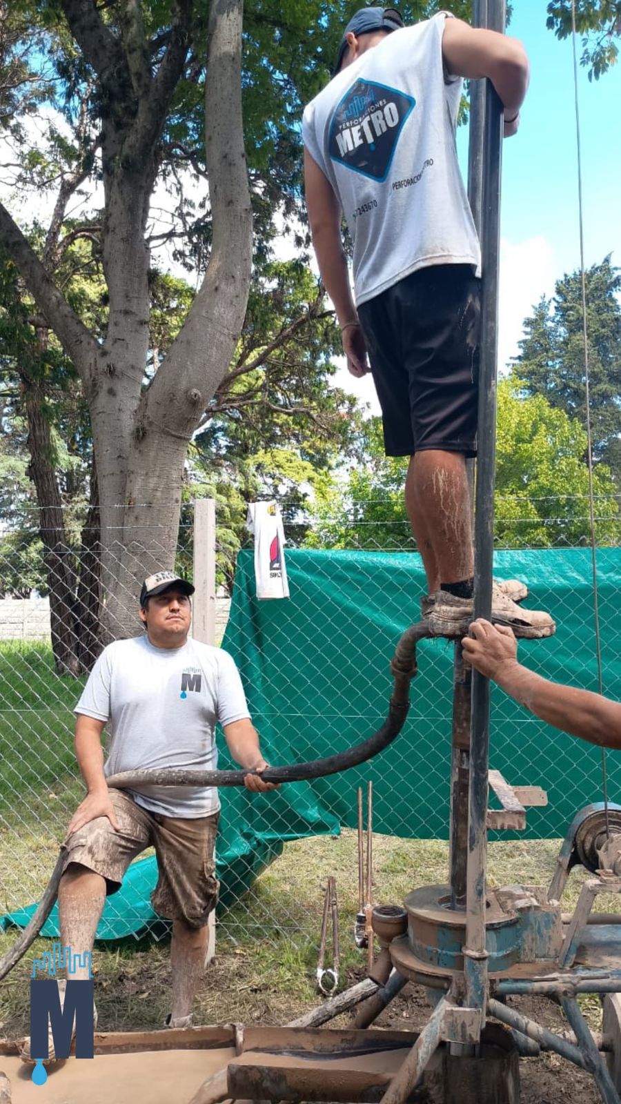 Trabajadores operando una perforadora de pozos al aire libre; un hombre está de pie sobre la plataforma y otro cerca.