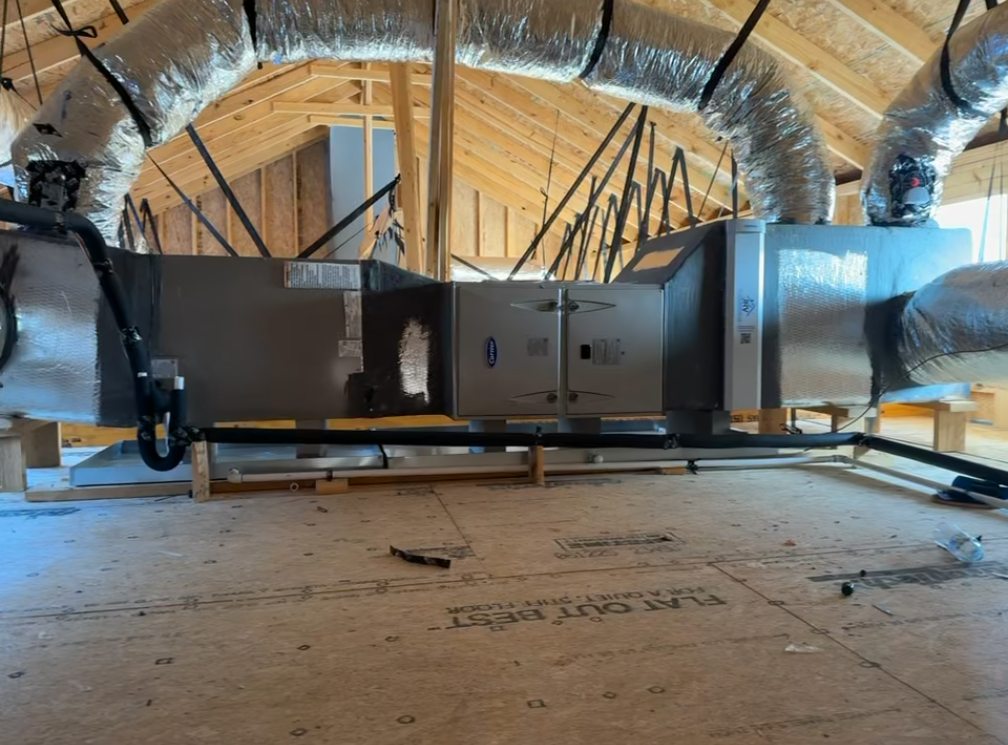 A central HVAC air handler unit and silver insulated ductwork installed in an attic with wooden trusses.