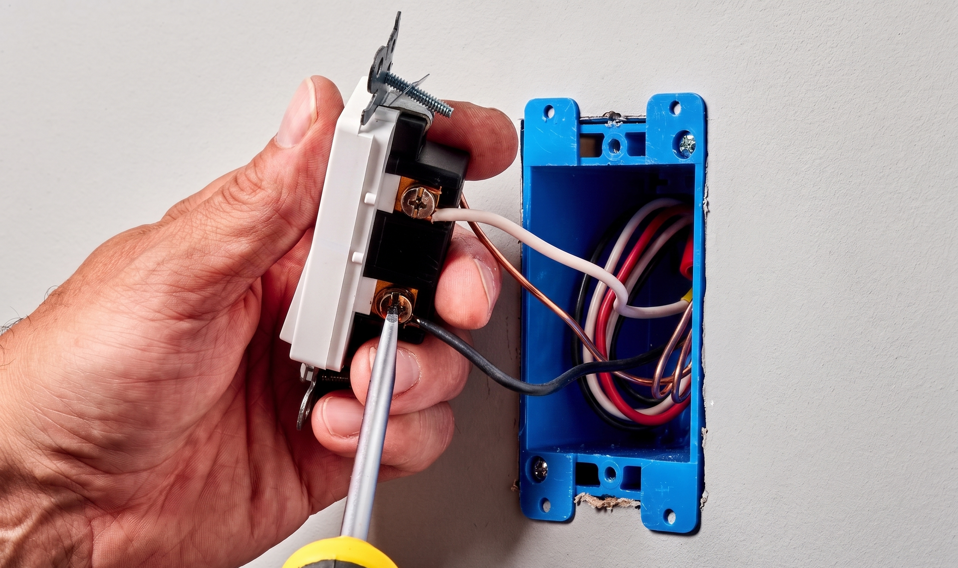 Hand wiring a white switch into a blue electrical box with a screwdriver nearby