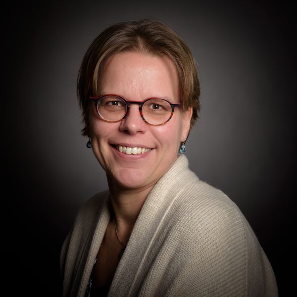 Een lachend persoon met kort haar, een bril en een lichtgekleurd vest, tegen een donkere, wazige achtergrond.