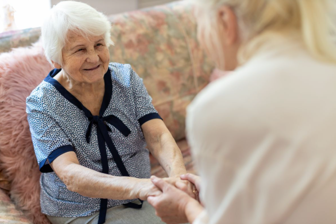 Een oudere man met wit haar glimlacht terwijl hij hand in hand zit met iemand die met zijn rug naar hem toe staat, binnenshuis op een bank.