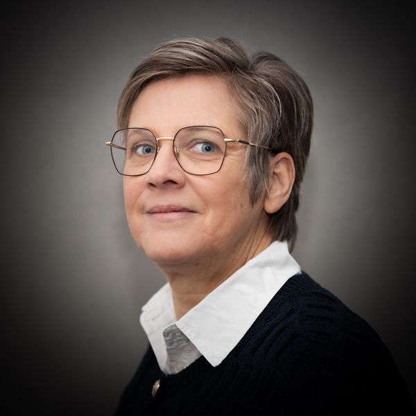 Portret van een persoon met een bril, een wit overhemd met kraag en een donkere trui, tegen een neutrale grijze achtergrond.