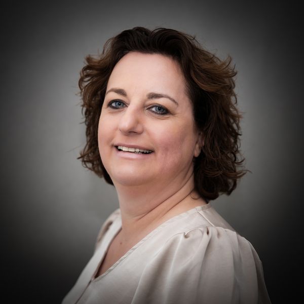 Professionele portretfoto van een lachend persoon met schouderlang, golvend bruin haar, gekleed in een lichtgekleurde top.