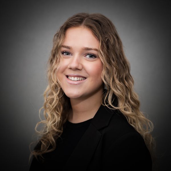 Portretfoto van een lachend persoon met schouderlang, golvend blond haar, gekleed in een zwarte blazer, tegen een grijze achtergrond.
