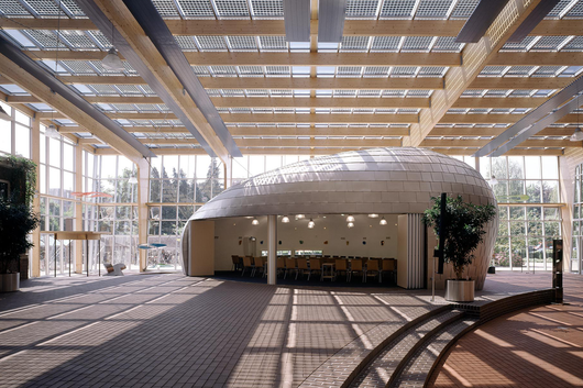 Een licht, modern atrium met een houten rasterplafond en een grote, ronde houten constructie met daarin een vergaderruimte.