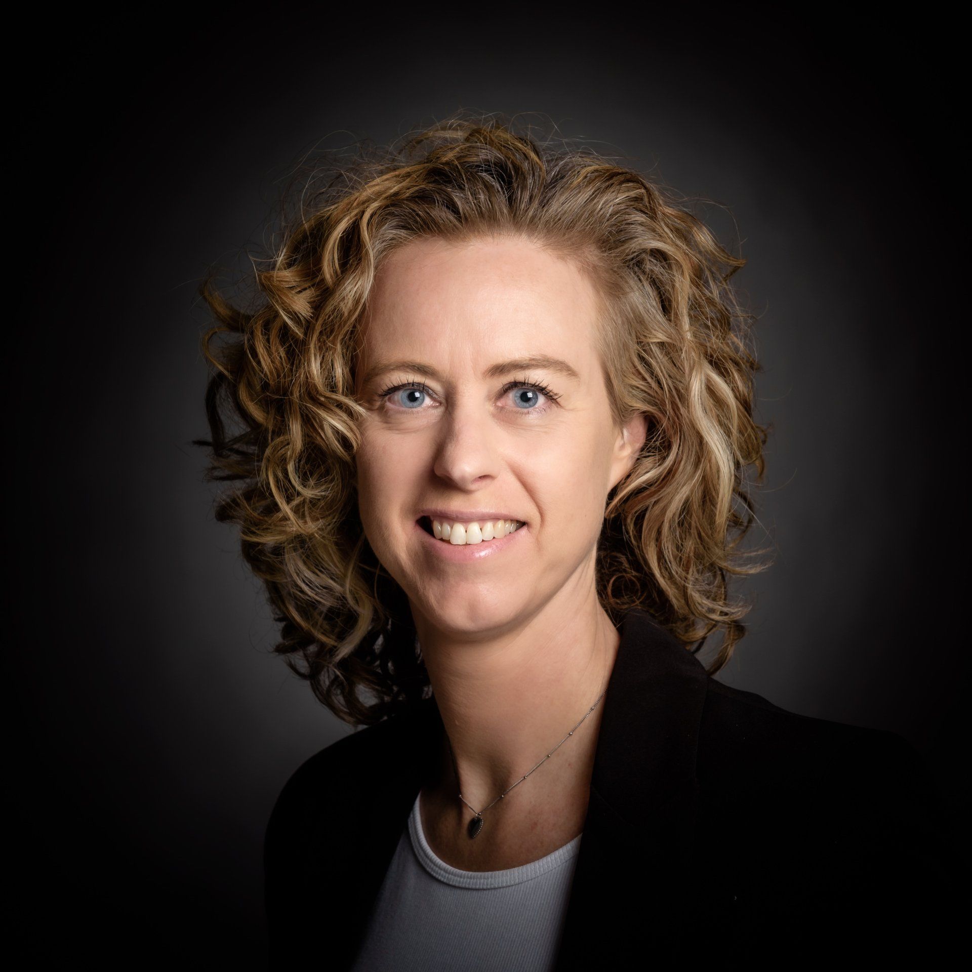 Een lachend persoon met krullend haar, gekleed in een wit overhemd en een donkere blazer, tegen een donkere, onscherpe achtergrond.