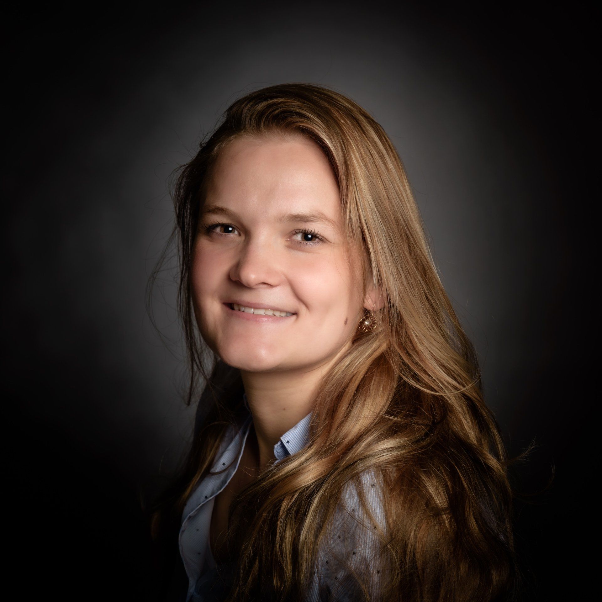 Een glimlachend persoon met lang, lichtbruin haar, gekleed in een lichtblauw overhemd met kraag, tegen een donkergrijze achtergrond.