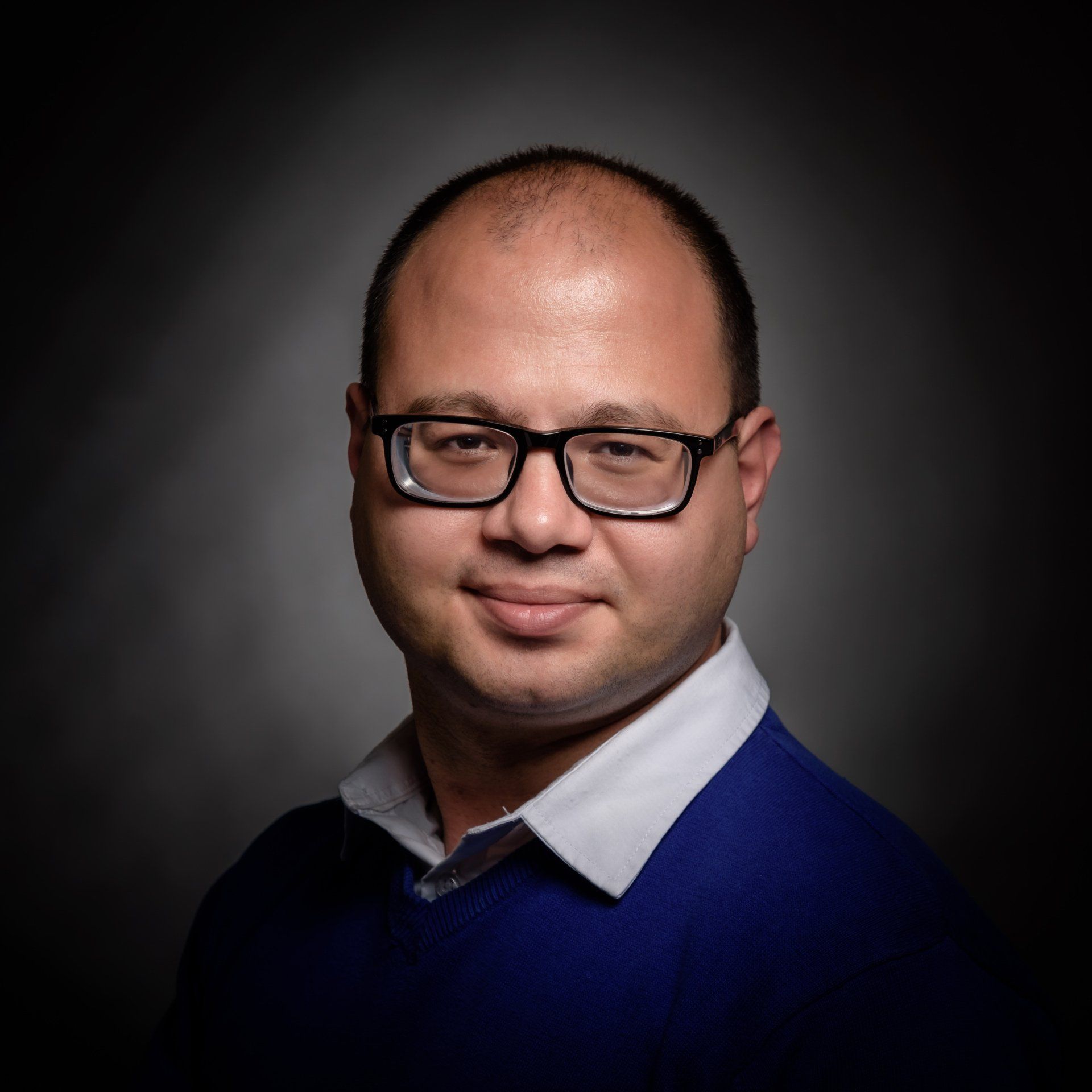 Een persoon met kort haar en een bril, gekleed in een blauwe trui en een overhemd met kraag, tegen een donkere, vignetachtige achtergrond.
