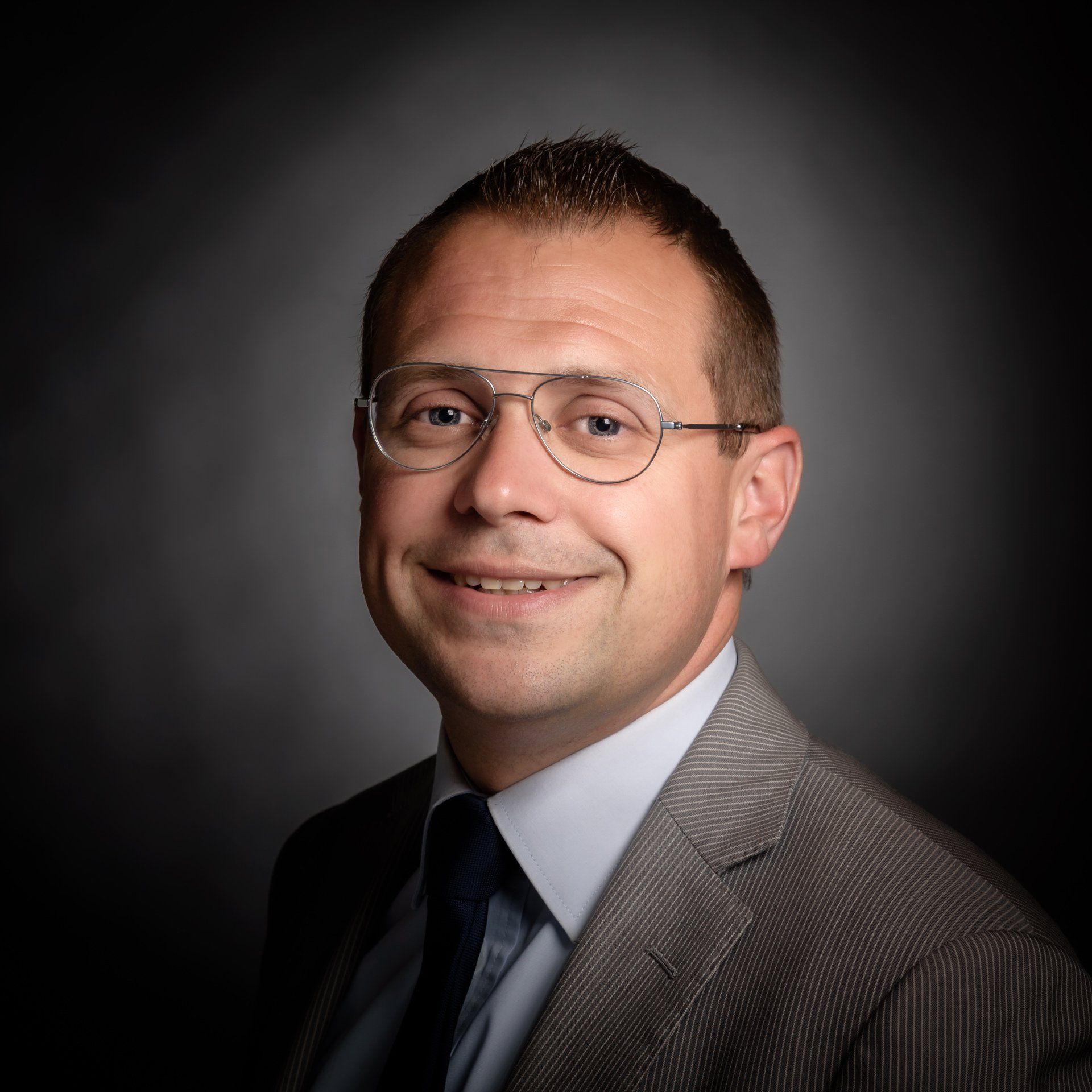 Een portretfoto van een persoon met een bril, een licht overhemd, een donkere stropdas en een colbert met patroon, tegen een donkere achtergrond.