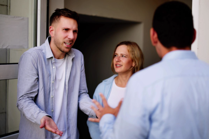 Drie mensen staan ​​bij een open deuropening en voeren een gesprek met levendige handgebaren en aandachtige gezichtsuitdrukkingen.