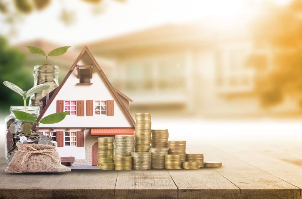 Een modelhuis met een klein plantje en een stapel gouden munten op een houten tafel, met een wazig huis op de achtergrond.