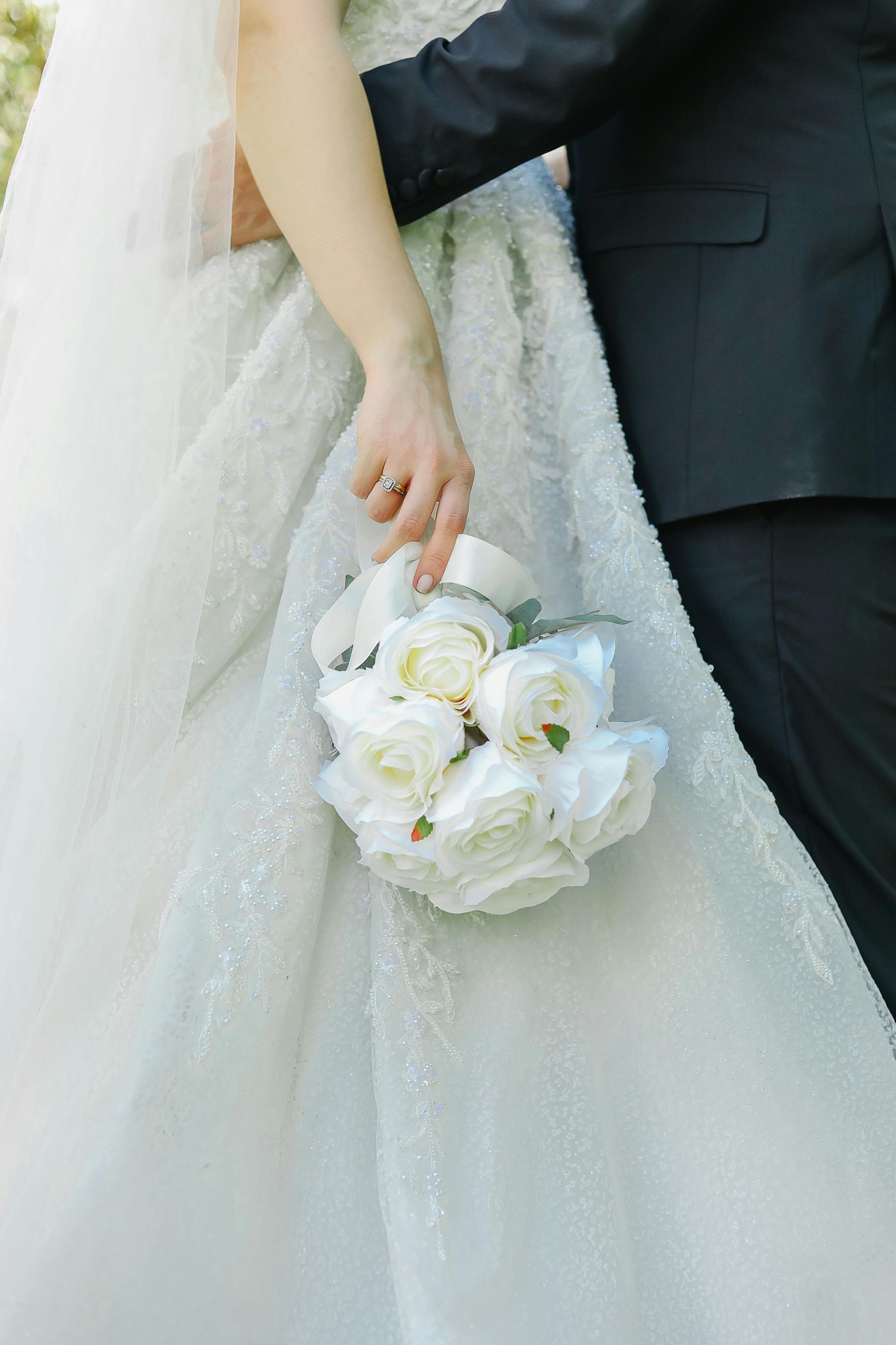 Een bruid in een witte jurk met textuur houdt een boeket witte rozen vast naast haar partner in een zwart pak.