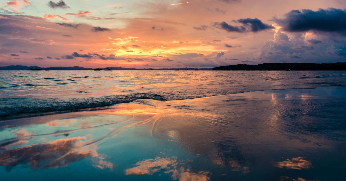 Een serene zonsondergang boven een kalme oceaan, met zachte wolken die weerspiegelen in het natte, glinsterende zand aan de kustlijn.