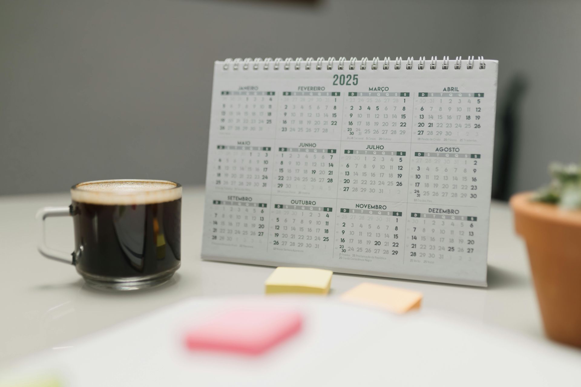 Op een witte tafel naast een klein potplantje staan een bureaukalender voor 2023, een mok zwarte koffie en wat plakbriefjes.