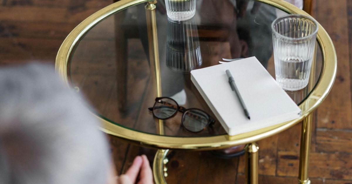 Op een tafel met een glazen blad liggen een notitieboekje, een pen, een bril en een glas water, van bovenaf gezien.
