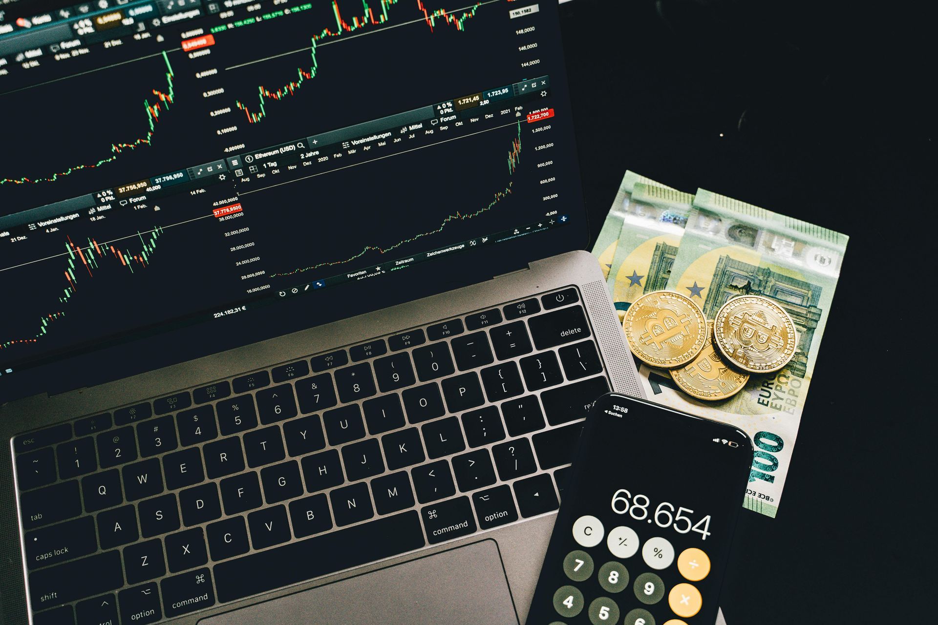 Een laptop met financiële aandelengrafieken, een rekenmachine en opgestapelde bankbiljetten op een donkere achtergrond.