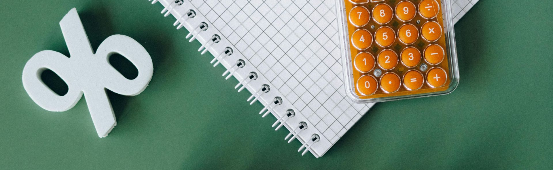 Een groot wit procentteken staat naast een notitieboekje met ruitjespapier en een kleine rekenmachine op een donkergroene achtergrond.
