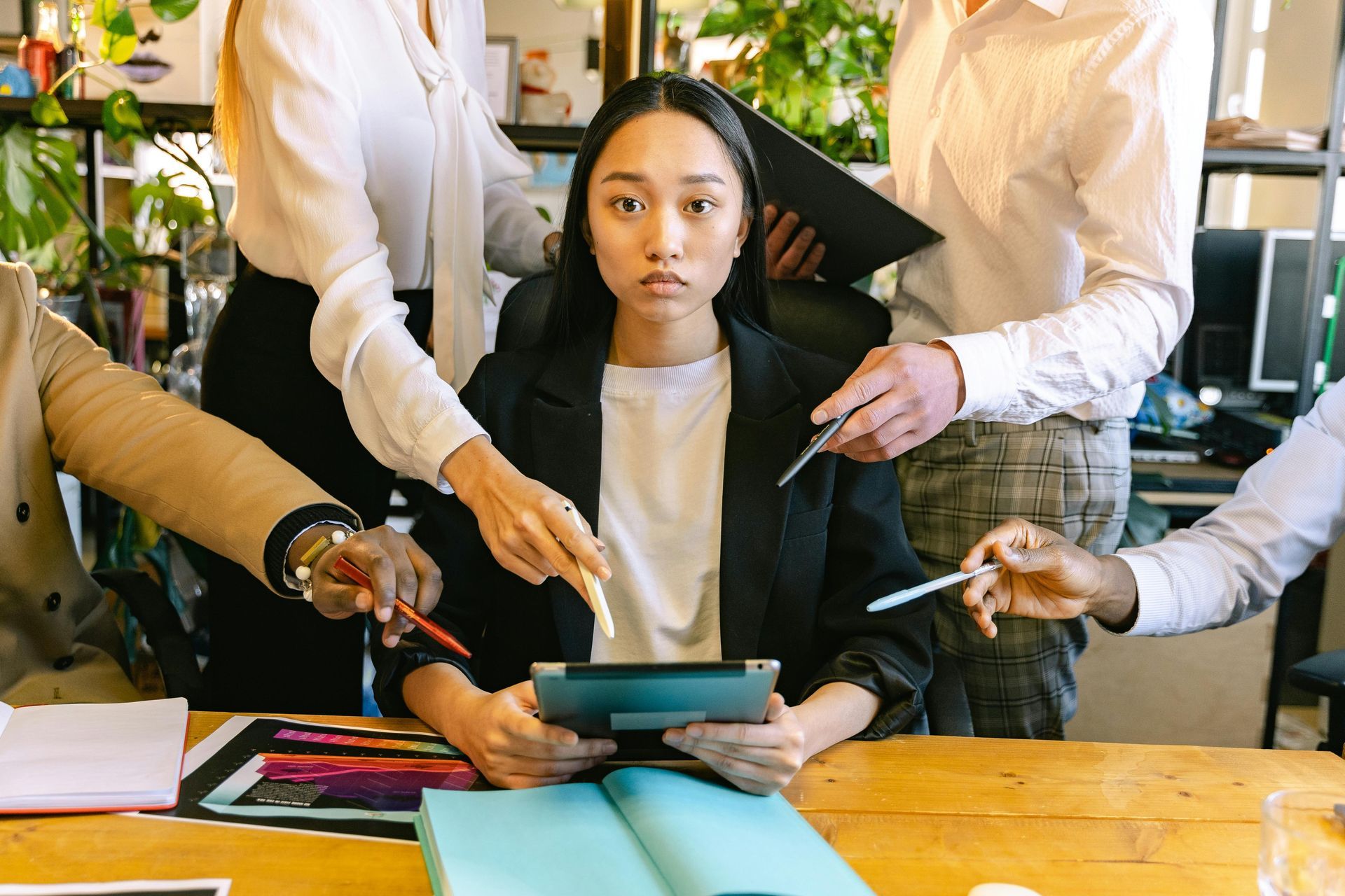 In een drukke kantooromgeving houdt een persoon in het midden een tablet vast, terwijl vanuit verschillende richtingen met pennen naar hem of haar wordt gewezen.