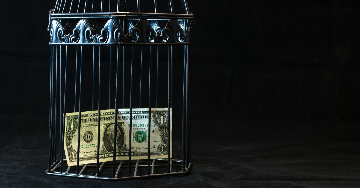 Een dollarbiljet zit gevangen in een zwarte, decoratieve vogelkooi tegen een donkere achtergrond.