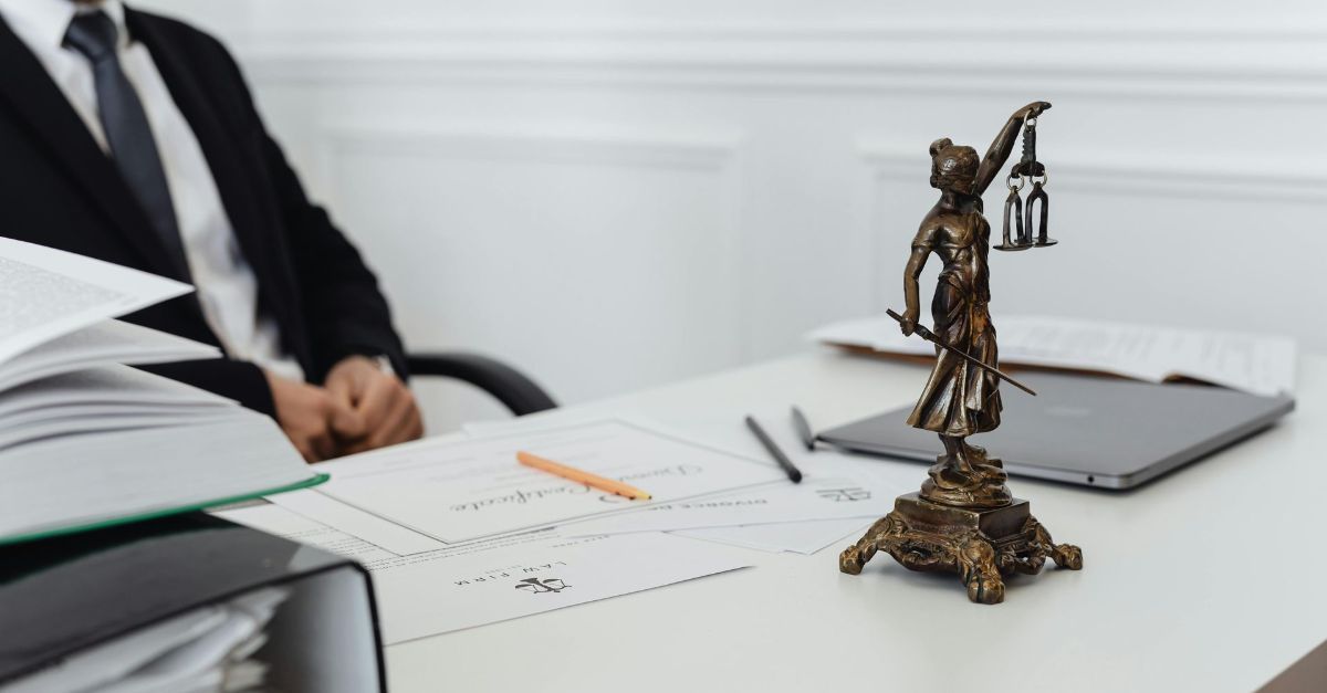 Een advocaat in pak zit aan een bureau met een bronzen beeld van Vrouwe Justitia, documenten en pennen in een lichte, professionele kantoorruimte.