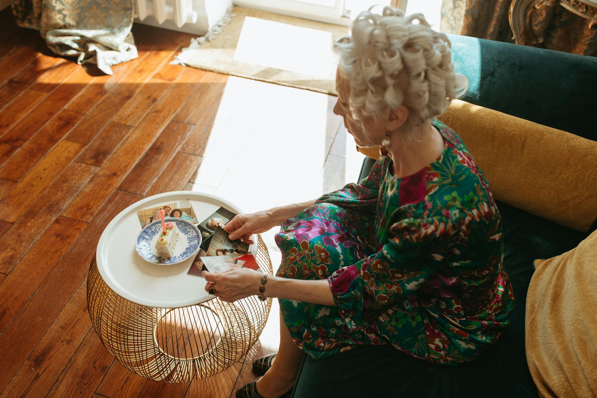 Een vrouw met gestyled haar en een bloemenjurk zit op een bank en reikt naar een klein taartje op een bijzettafel.
