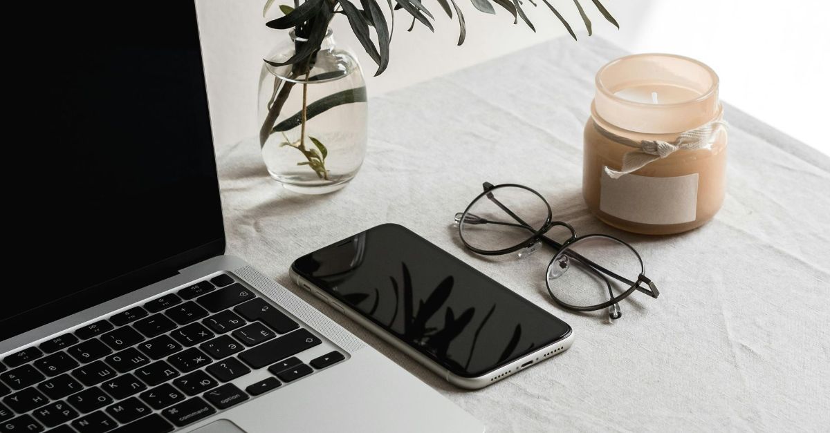 Een laptop, smartphone, bril, een klein kaarsje en een plant in een glazen vaas staan uitgestald op een witte tafel.