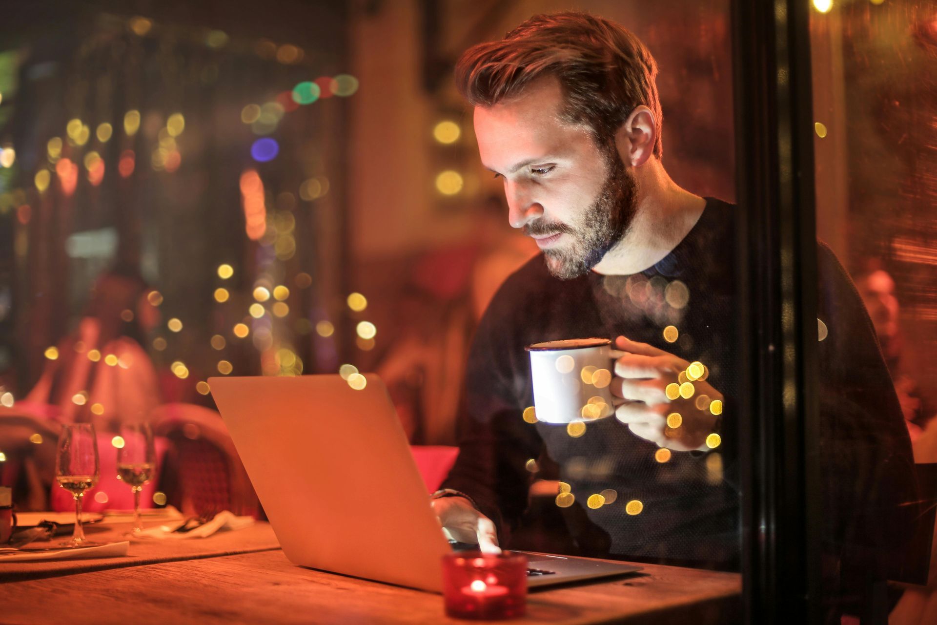 Een bebaarde man in een donkere trui zit aan een tafel met een laptop en een mok in zijn hand, verlicht door warme caféverlichting.