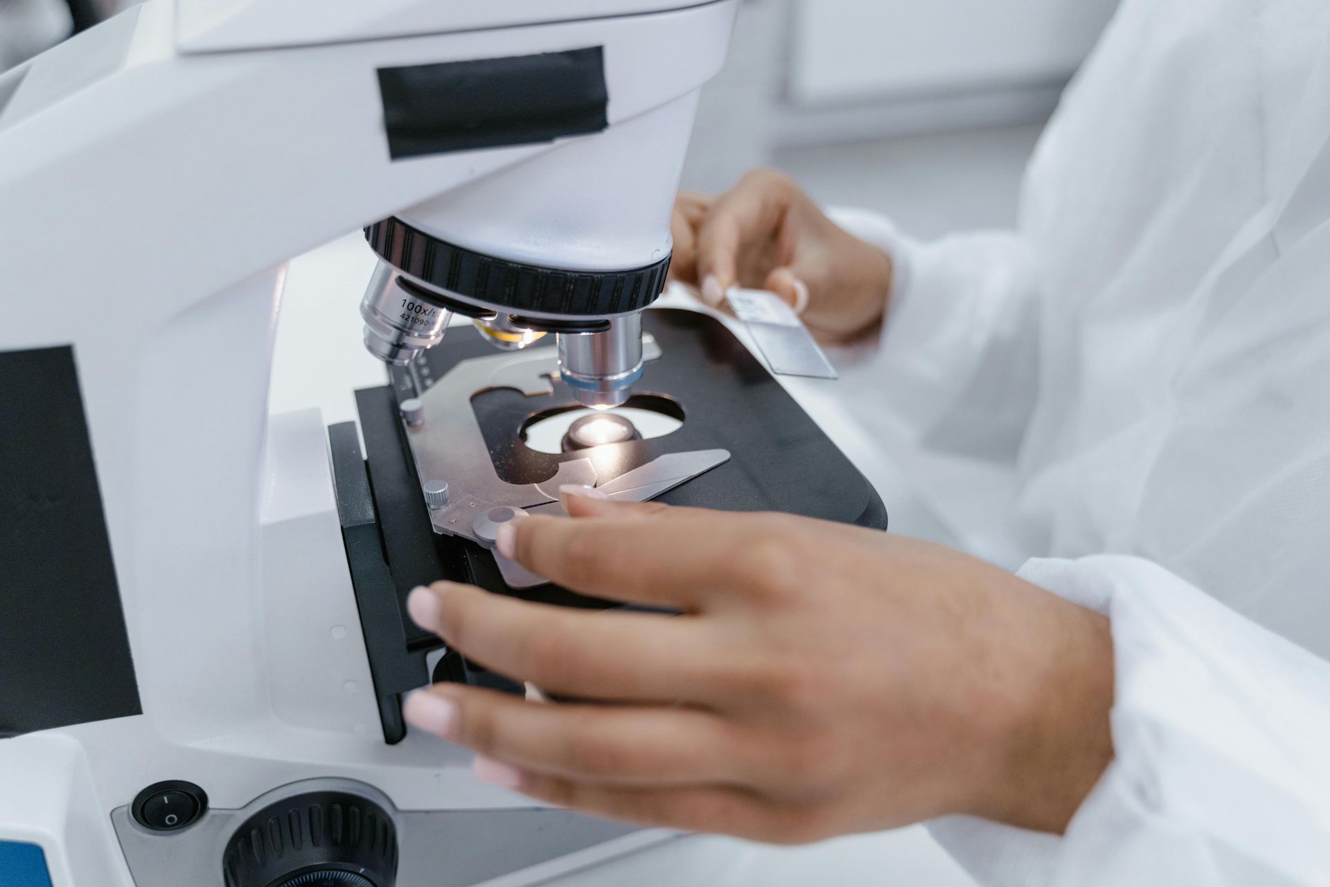 Een wetenschapper in een witte laboratoriumjas plaatst een objectglaasje op de tafel van een laboratoriummicroscoop.