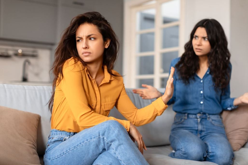 Twee mensen zitten op een bank in de woonkamer; de een kijkt weg terwijl de ander gebaren maakt tijdens een gespannen gesprek.