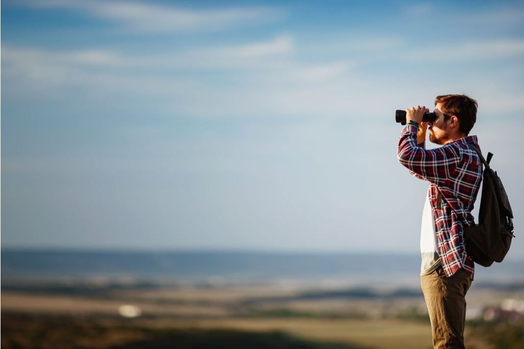 Een wandelaar, gekleed in een geruit shirt en met een rugzak, staat op een heuvel en kijkt door een verrekijker naar een wazig landschap.