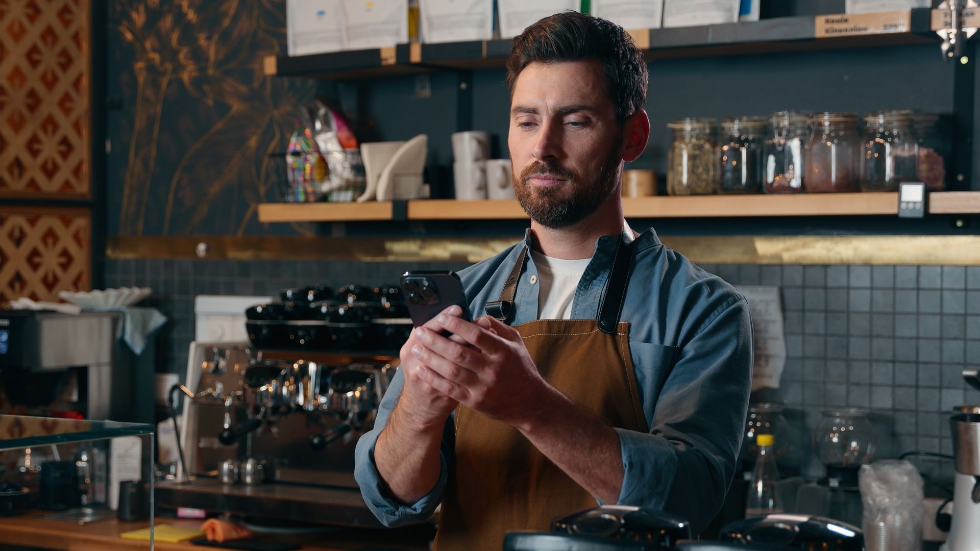 Barista in a café, looking down while holding a cup, with coffee equipment and shelves behind him