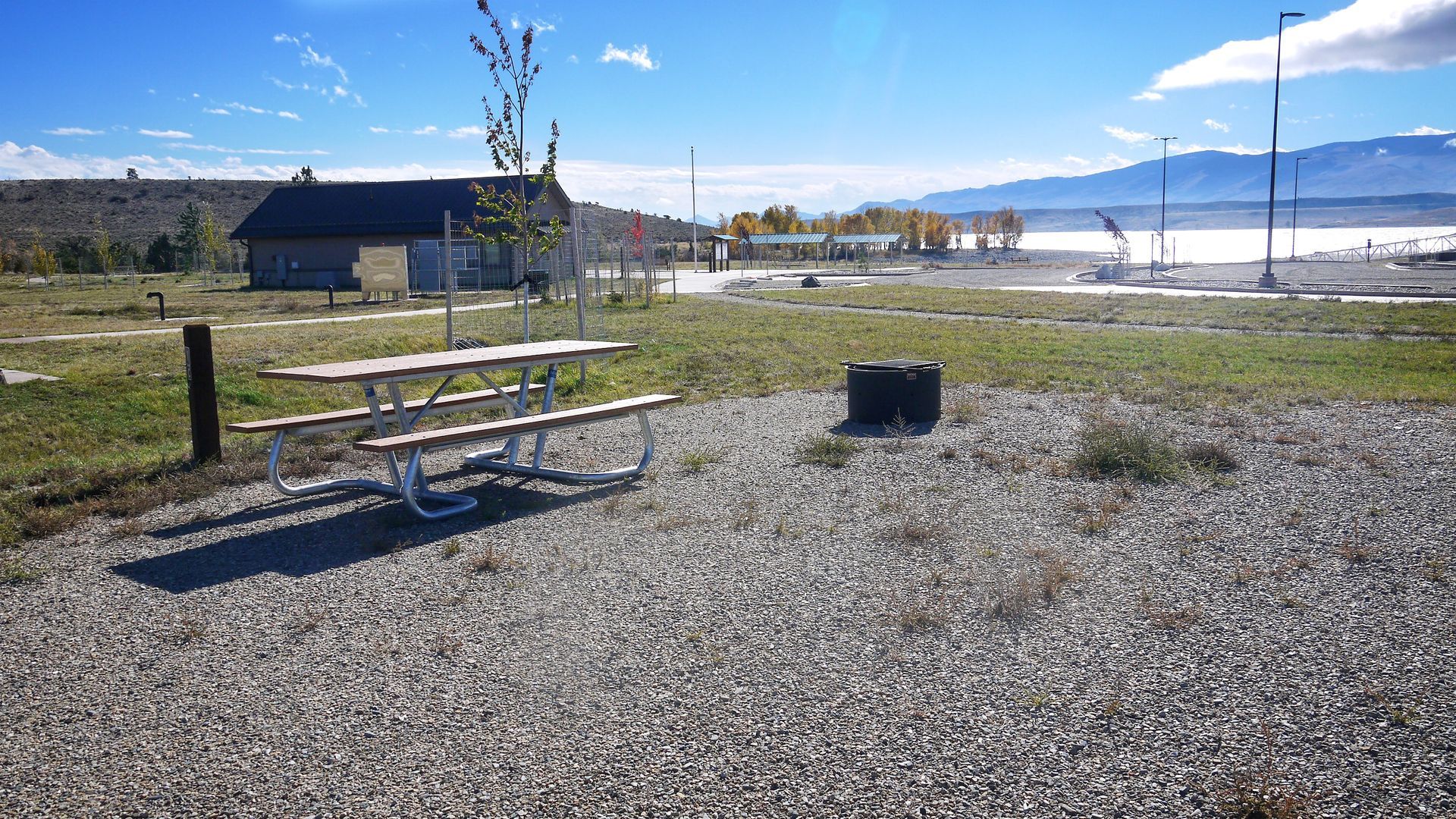 Picnic table beside a gravel campsite with mountains and a lake in the background under a blue sky